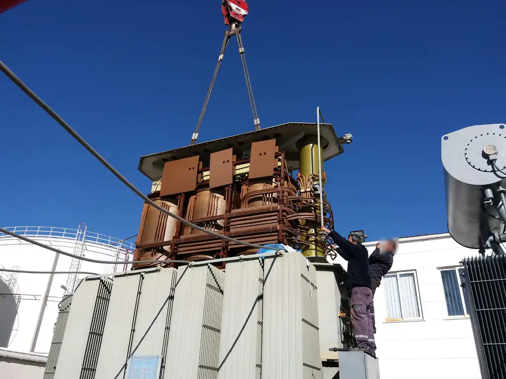 A transformer active part hanging from a crane hook above its open tank against a deep blue sky, with two fitters at the tank edge