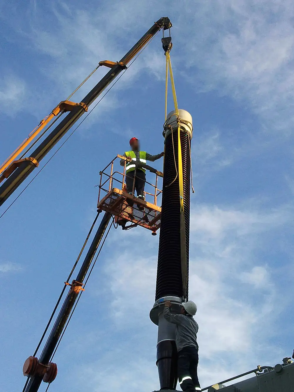 A technician in a crane-mounted basket guiding a tall high-voltage bushing on a lifting sling, while a second technician steadies its base at the transformer