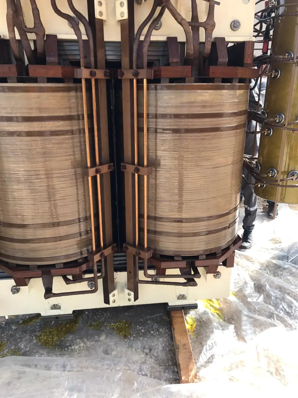 Two wound limbs of a transformer active part with their clamping structure and copper leads, standing on plastic sheeting outdoors, a tap-changer cylinder at the right