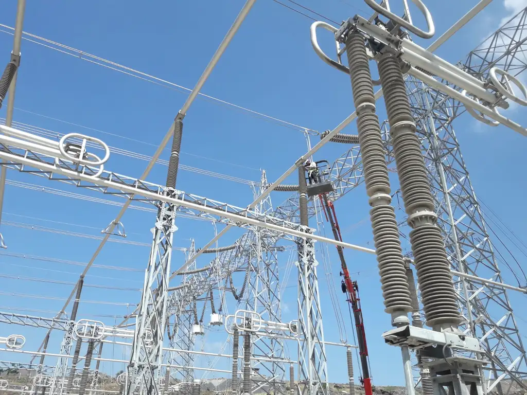 Looking up at high-voltage disconnectors and tall ribbed insulator columns on lattice steel gantries, with a technician working from a mobile elevating work platform against a blue sky
