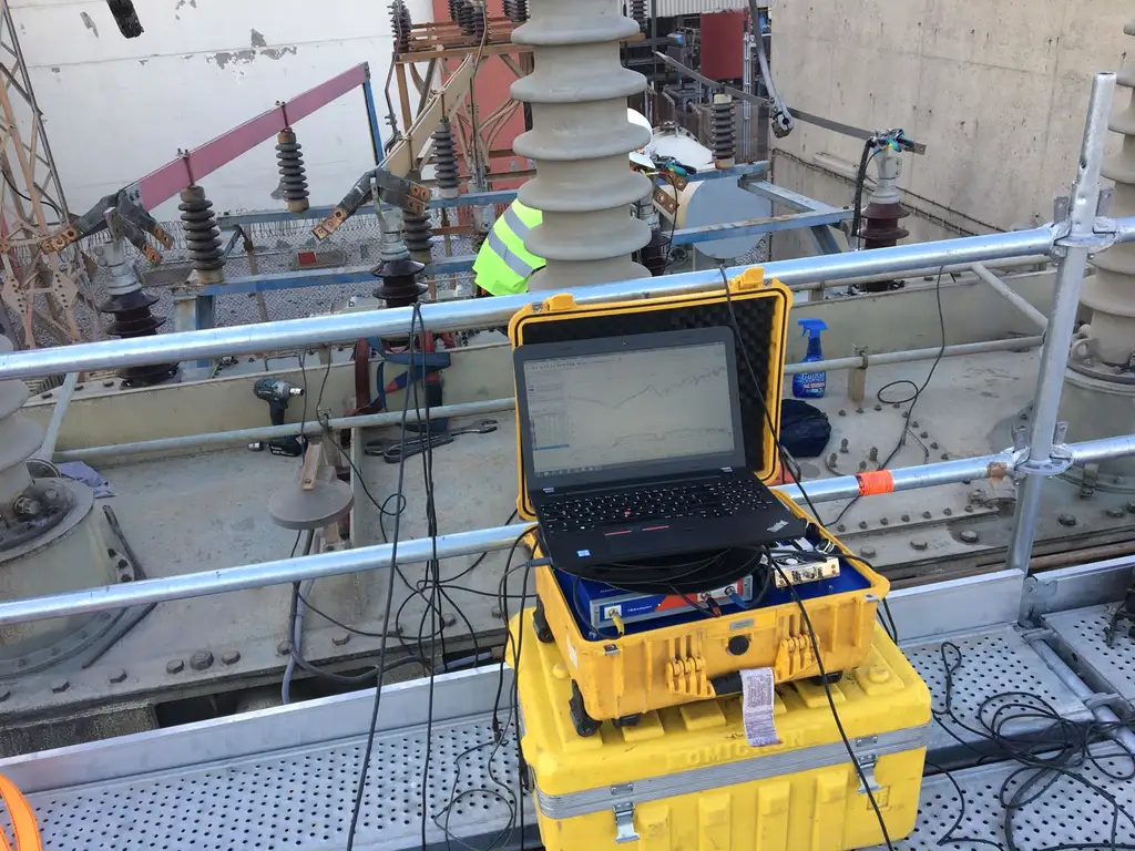 A laptop showing frequency-response traces, set on the open case of a test instrument on a transformer cover, with test leads running to the bushings