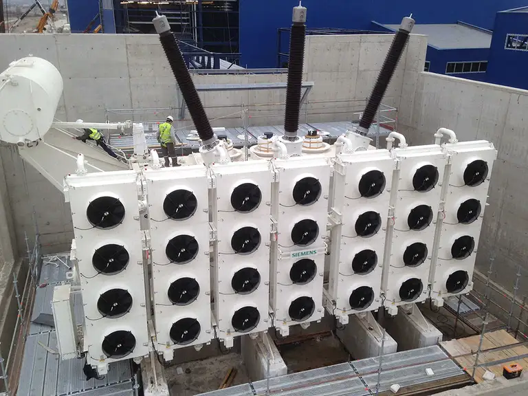 A large power transformer in a concrete pit, with three high-voltage bushings above a bank of fan-cooled oil coolers and two technicians in hi-vis vests working on the tank cover