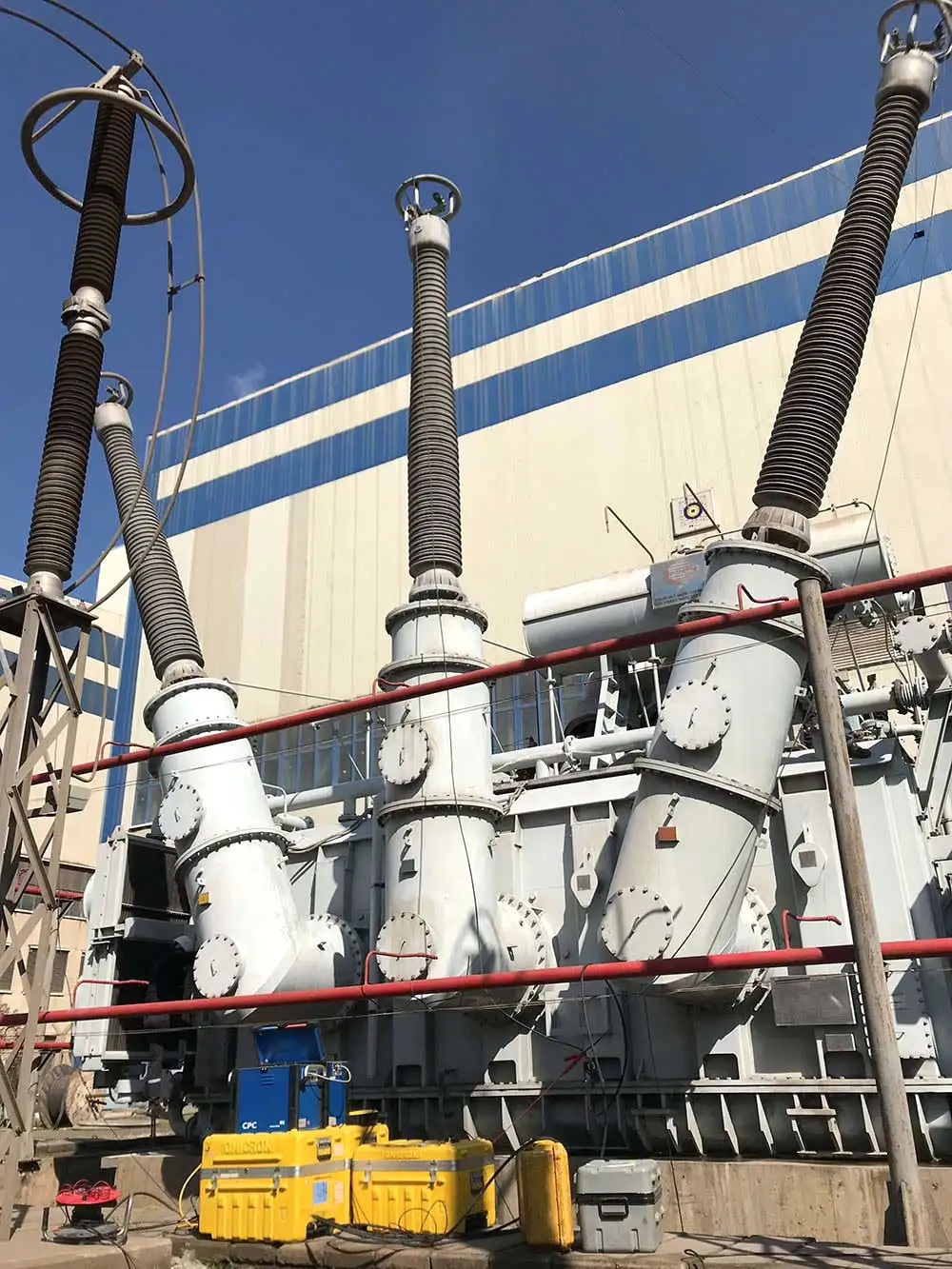 A large power transformer with three tall high-voltage bushings on angled turrets, and a portable test set with its yellow transport cases set down on the plinth at its base