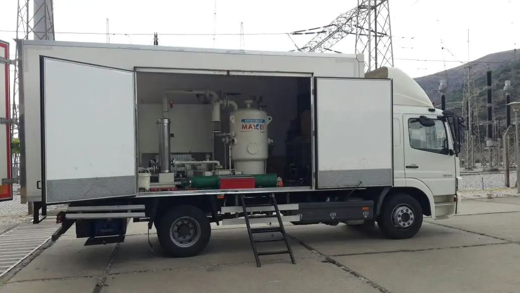 A box-body truck with its side doors open, showing a vacuum oil-treatment plant fitted inside, parked on a concrete apron beside a switchyard with hills behind