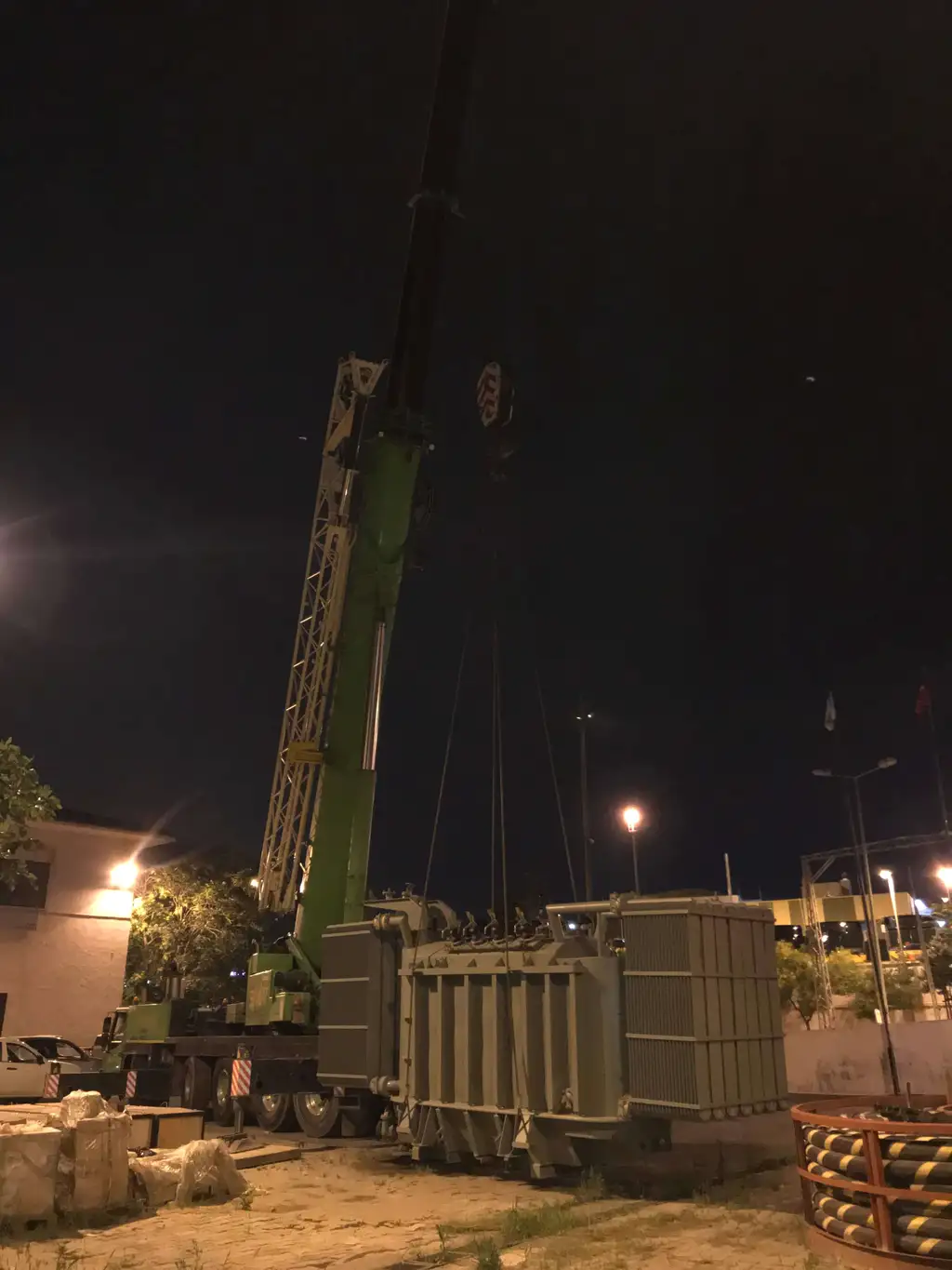 A power transformer rigged to the hook of a truck-mounted crane at night, lifting slings taut under street lighting, with wrapped crates and a cable drum in the foreground