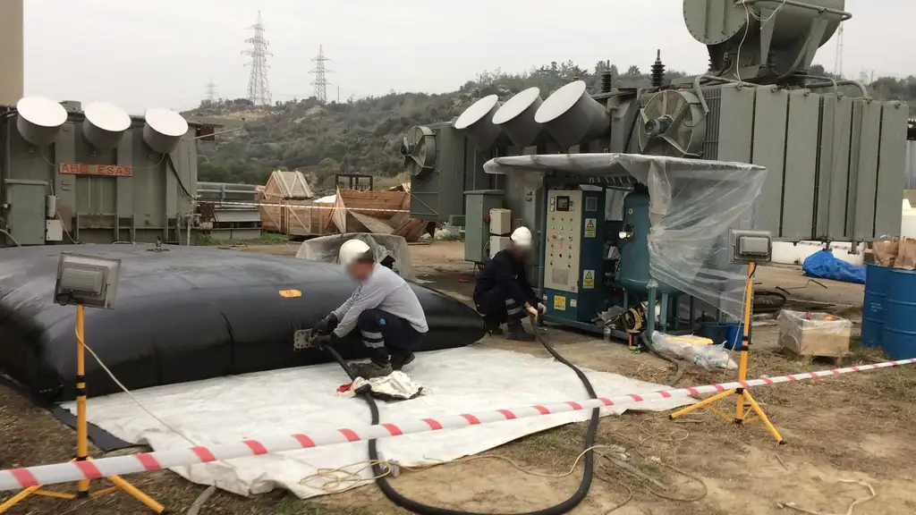 Two technicians connecting hoses between a large black oil storage bladder and a treatment plant under a plastic sheet, beside a power transformer, the work area taped off