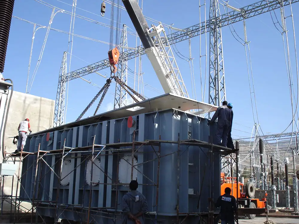 A mobile crane lifting the cover from a large transformer tank on chains in an outdoor switchyard, fitters on the tank top and on scaffolding around it